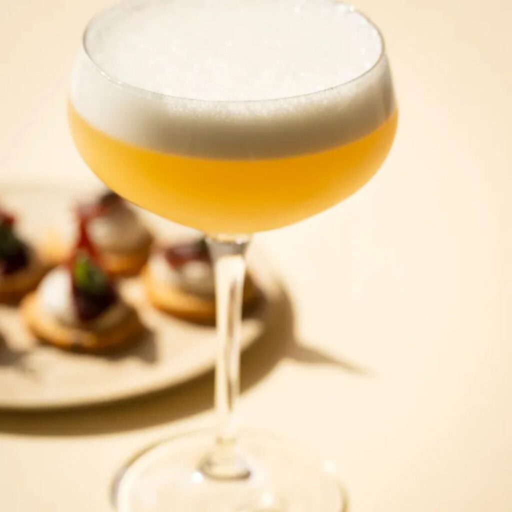 Bourbon-truffel sour met schuimlaagje en hapjes op een bord op de achtergrond