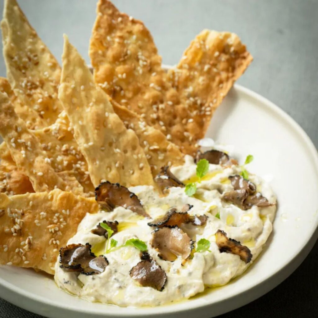 Romige hummus-truffeldip met truffelschijfjes, verse kruiden en crackers op een wit bord