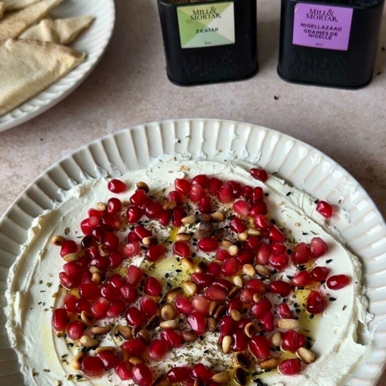 Bord met romige labneh dip, royaal bestrooid met rode granaatappelpitjes, geroosterde pijnboompitten, groene za’atar en zwarte nigellazaadjes, geserveerd met Libanees flatbread op de achtergrond.